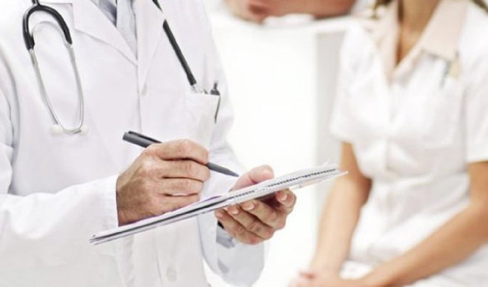 Bir doktorun klinik ortamda not aldığı ve arka planda hastanın görüldüğü sağlık danışmanlığı görseli.