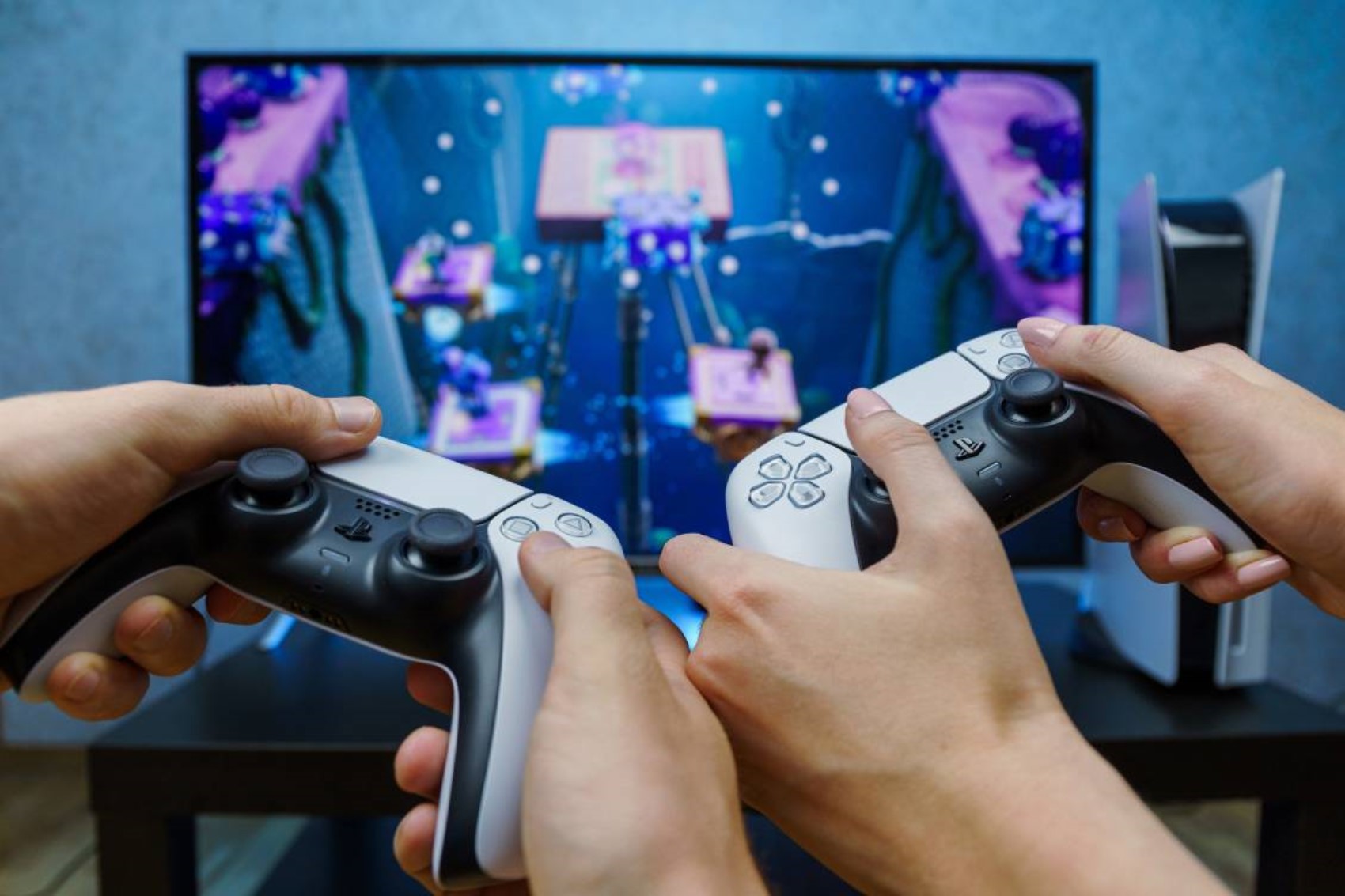 İki kişinin PlayStation kontrolcüleriyle televizyon karşısında oyun oynadığı görsel.