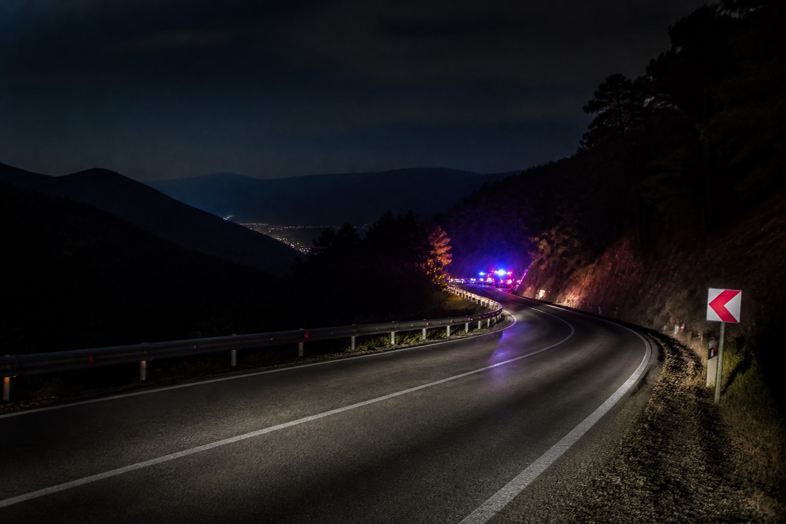 Gece vakti virajlı bir dağ yolunda bariyerler ve uzakta acil durum ışıkları görülen temsili görsel.