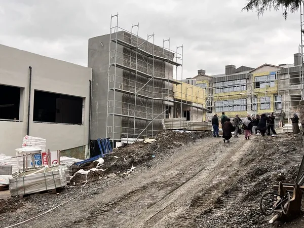 Yağışlı havada çamurlu zemine sahip okul inşaatı alanı ve bina cephesi görülüyor.