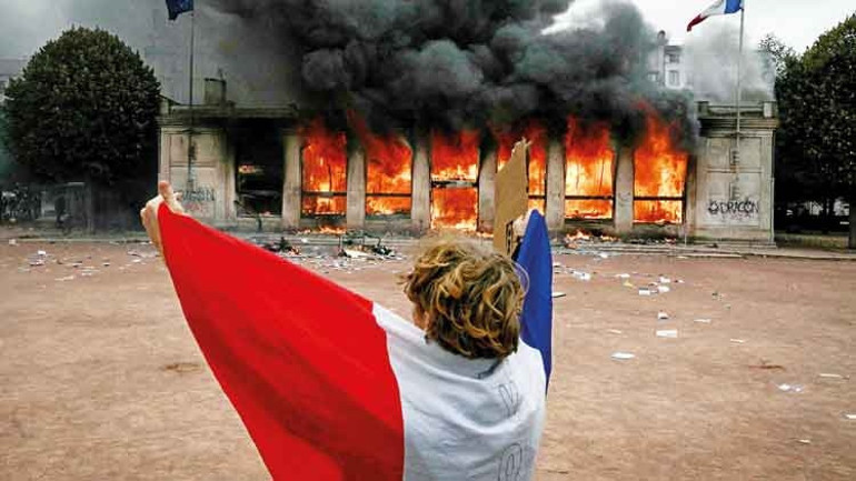 A protest scene where a person carrying a French flag stands in front of a burning building.