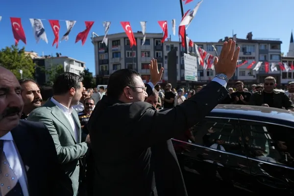 Kalabalık bir alanda araç yanında halka el sallayan takım elbiseli yerel yönetici.