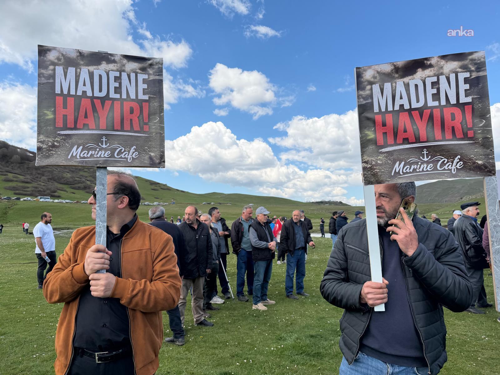 Çimenlik alanda madene karşı döviz taşıyan iki kişi ve arkada kalabalık.