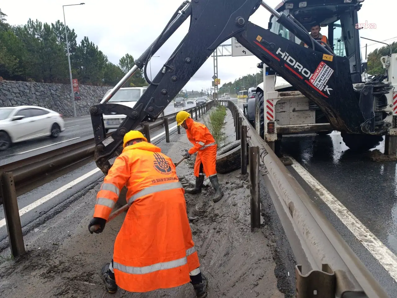 Turuncu kıyafetli belediye ekiplerinin yol kenarında çamur temizliği yaptığı görülüyor.