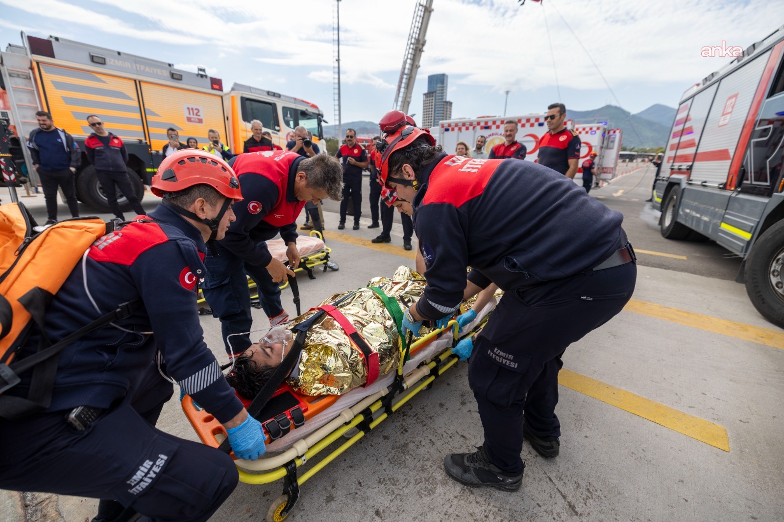 Drill footage showing fire crews carrying a casualty wrapped in a thermal blanket on a stretcher to an ambulance.