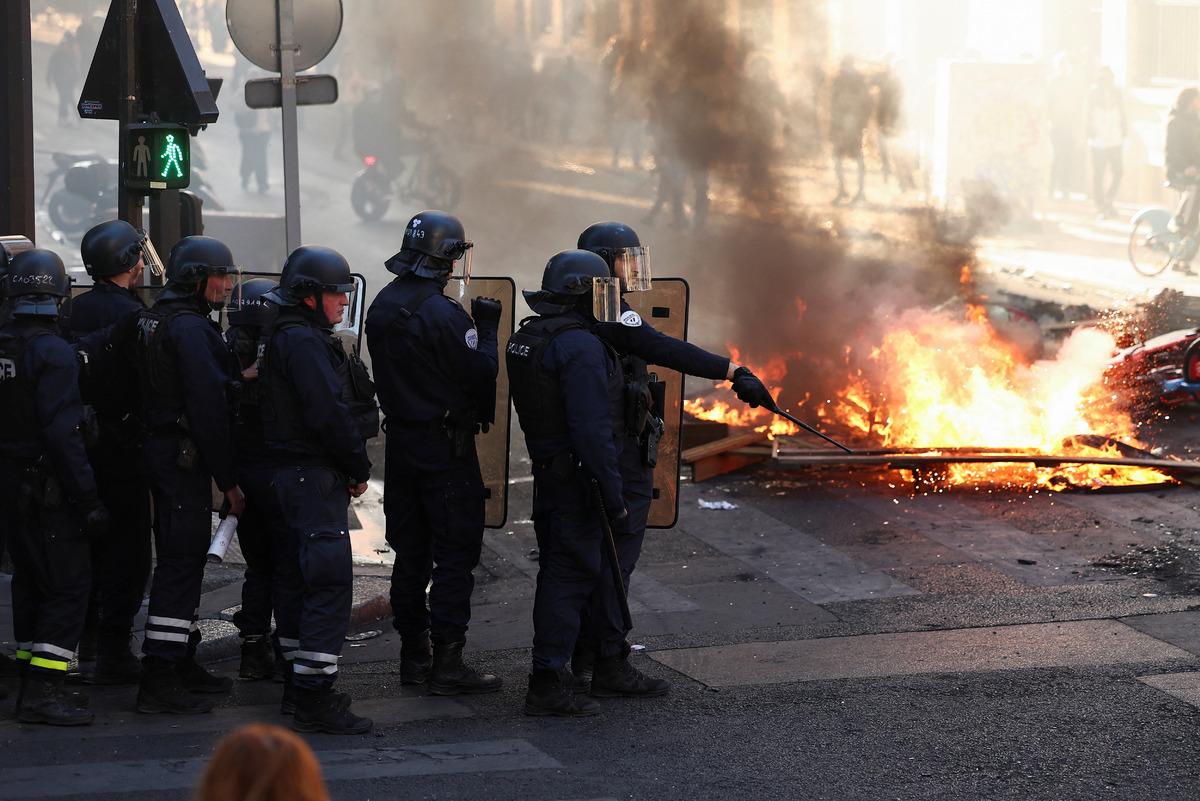 An image of riot police waiting on a street with burning barricades.