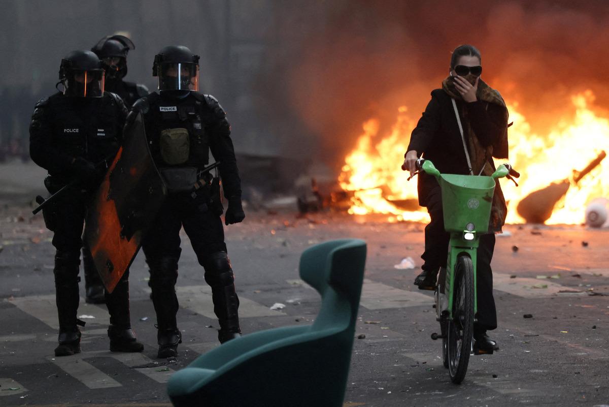 A street scene where police and a person on a bicycle are visible while flames burn in the background.
