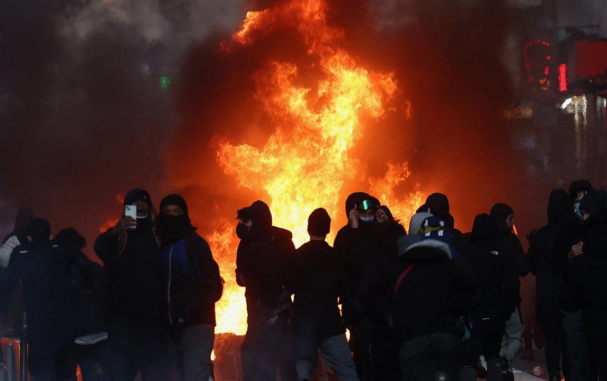 Silhouettes of masked protesters gathered in front of a large flame.