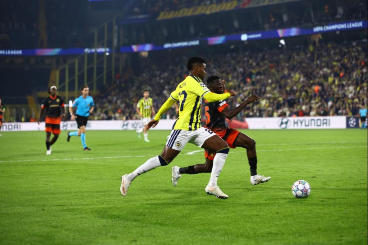 Fenerbahçe ve Slavia Prag oyuncularının top için koştuğu, arka planda tribünlerin yer aldığı maç fotoğrafı.
