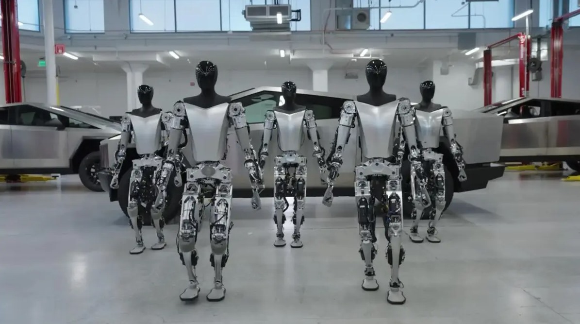 Tesla atölyesinde yan yana duran Optimus insansı robotları ve arkada elektrikli araçlar.