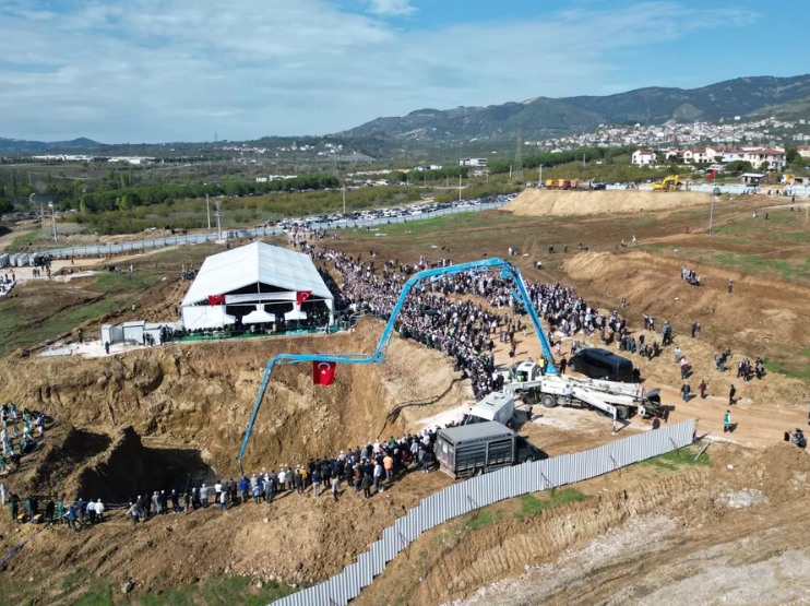 Geniş bir arazide kurulan beyaz tören çadırı, kalabalık katılımcılar, iş makineleri ve çevredeki yerleşim alanları görülüyor.