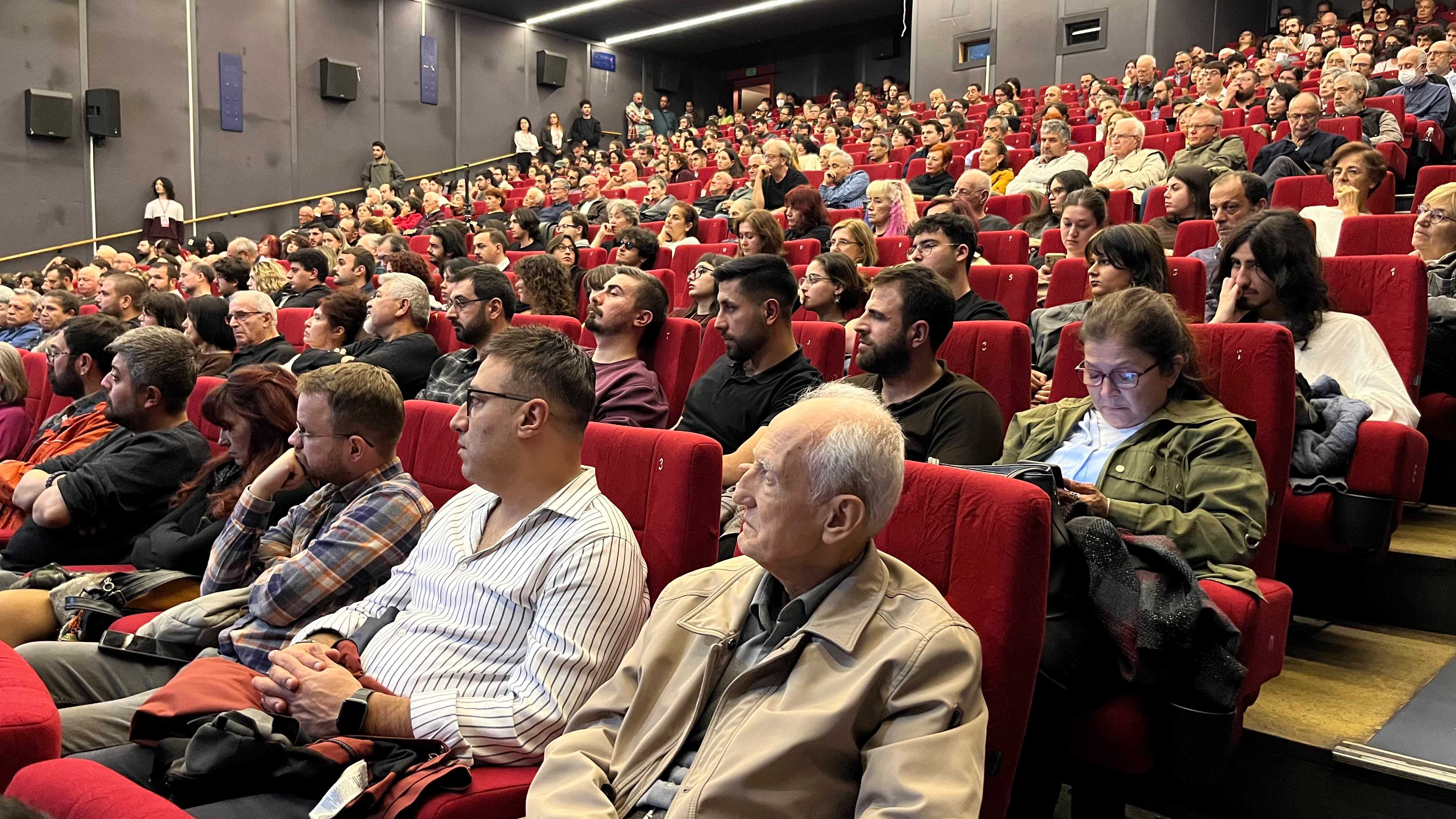 Kırmızı koltuklu konferans salonunda forumu takip eden kalabalık izleyiciler.