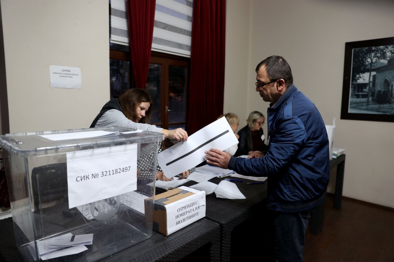 Seçmenin sandık görevlisine zarf uzattığı, şeffaf sandık ve seçim evraklarının bulunduğu bir oy verme alanı.