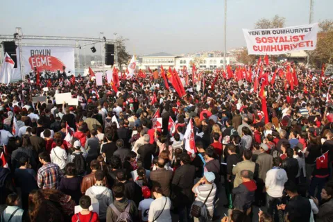 Kalabalık bir miting alanında kırmızı bayraklar taşıyan yurttaşlar ve Cumhuriyet temalı pankartlar görülüyor.