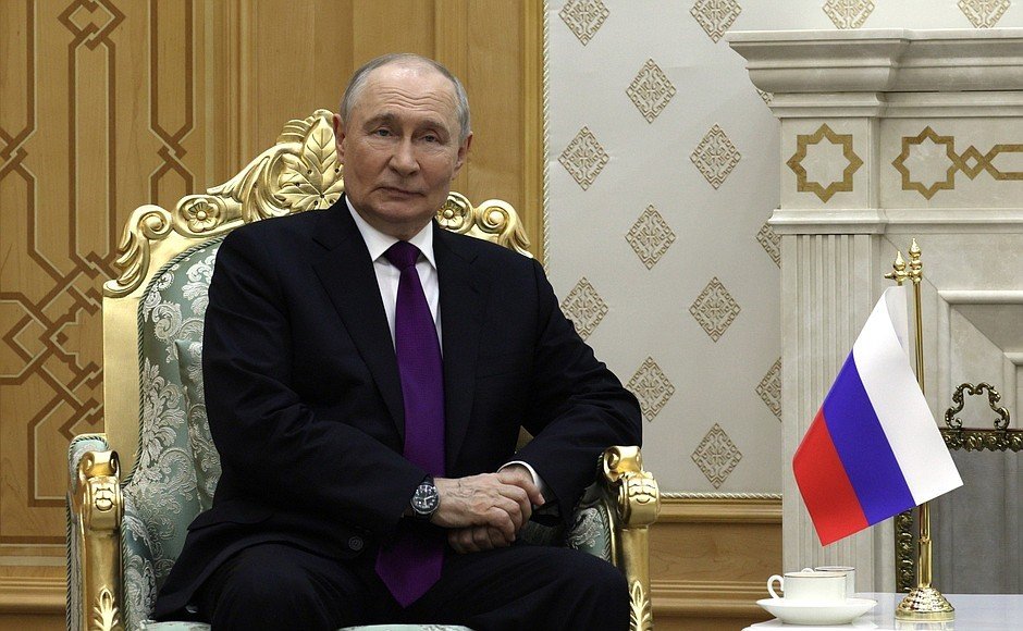 Wladimir Putin sitzt in einem offiziellen Saal mit russischer Flagge.