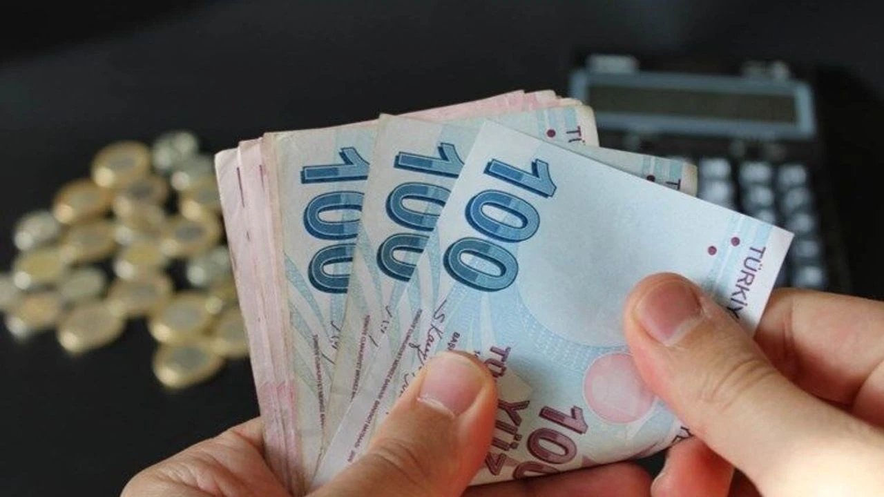 Elde tutulan Türk lirası banknotları ve arka planda madeni paraların yer aldığı görsel.