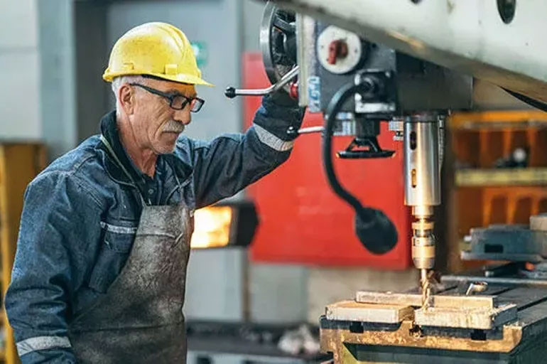 Atölyede makine başında çalışan kasklı yaşlı bir işçi.