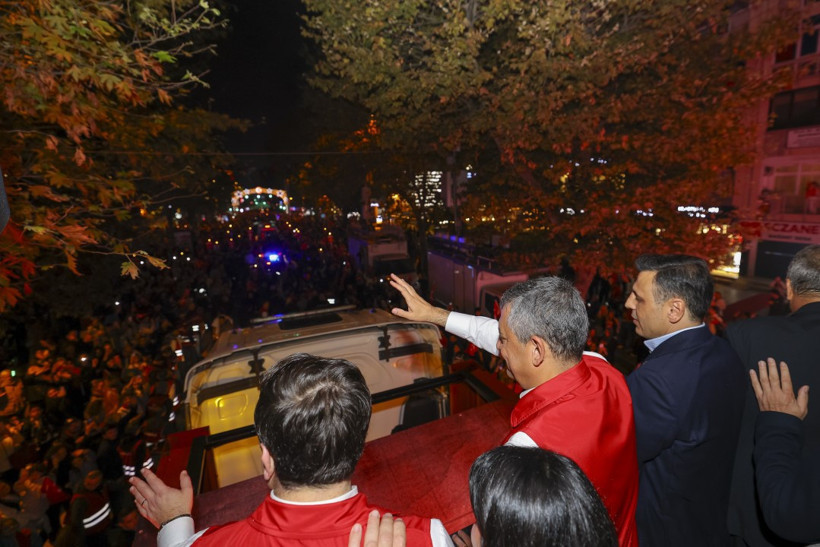 Özgür Özel’in aracın üzerinden kalabalığa el salladığı, ağaçlarla çevrili bir cadde görüntüsü.