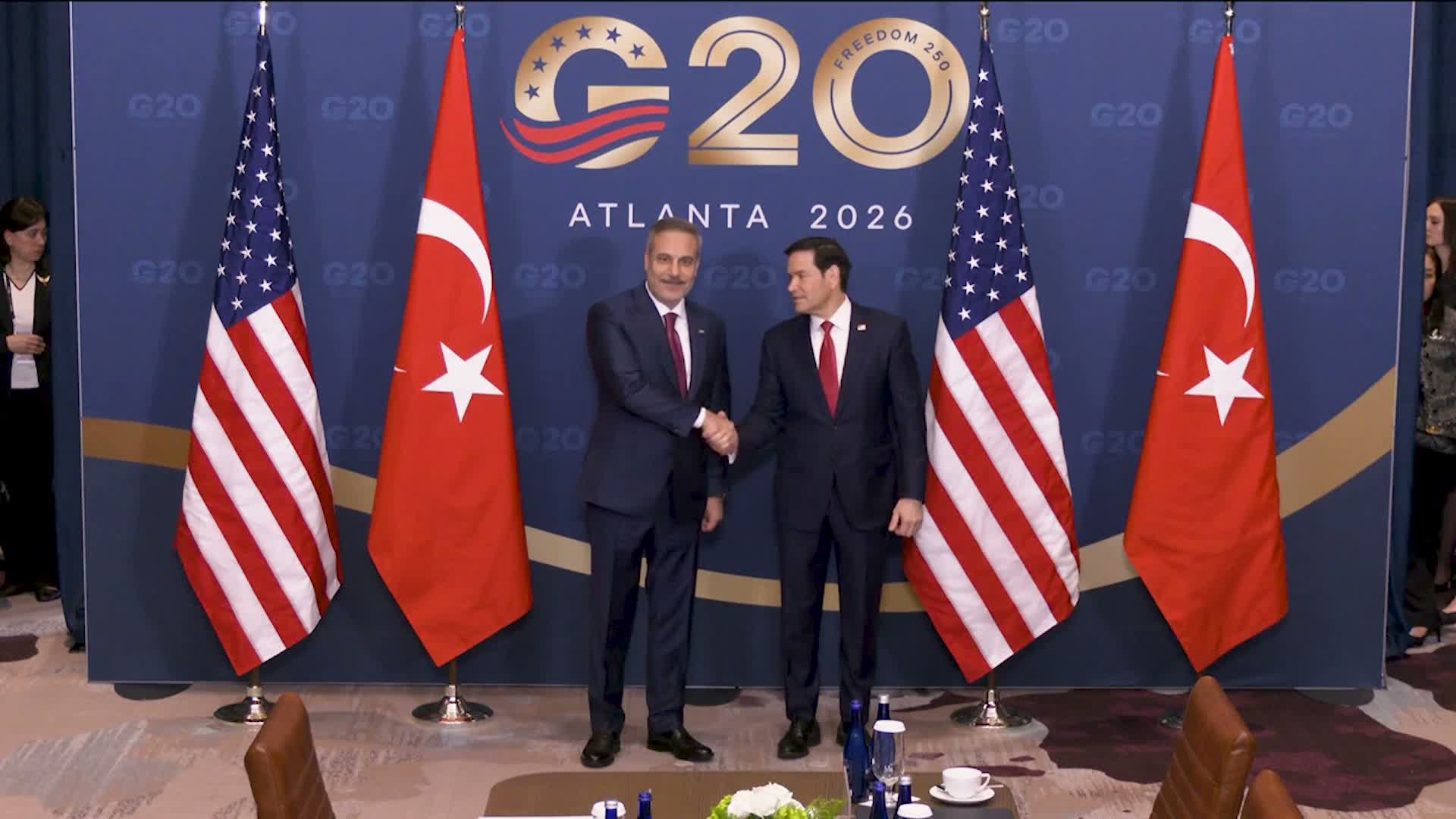 Hakan Fidan ve Marco Rubio, toplantı salonunda Türk ve ABD bayrakları önünde tokalaşıyor.