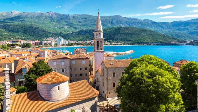 Budva’da deniz, tarihi yapılar, kilise kulesi ve dağ manzarası.