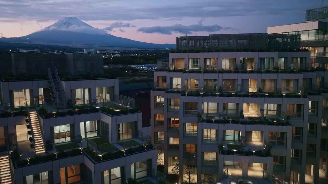 Akşam saatlerinde ışıkları yanan konaklama blokları ve uzakta dağ manzarası.