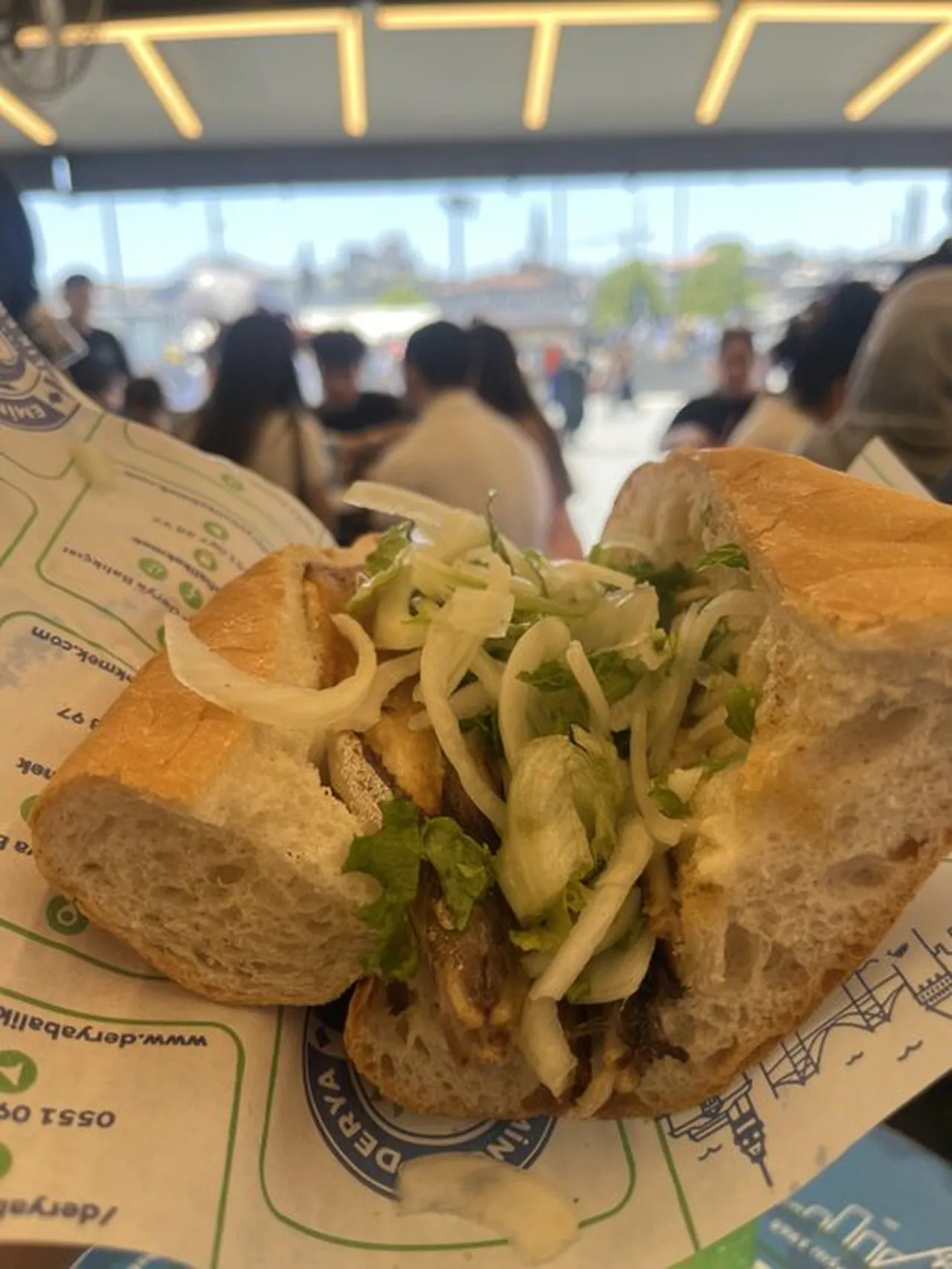 İstanbul’da bir mekanda servis edilen uskumru sandviçi ve arka planda kalabalık restoran görünümü.