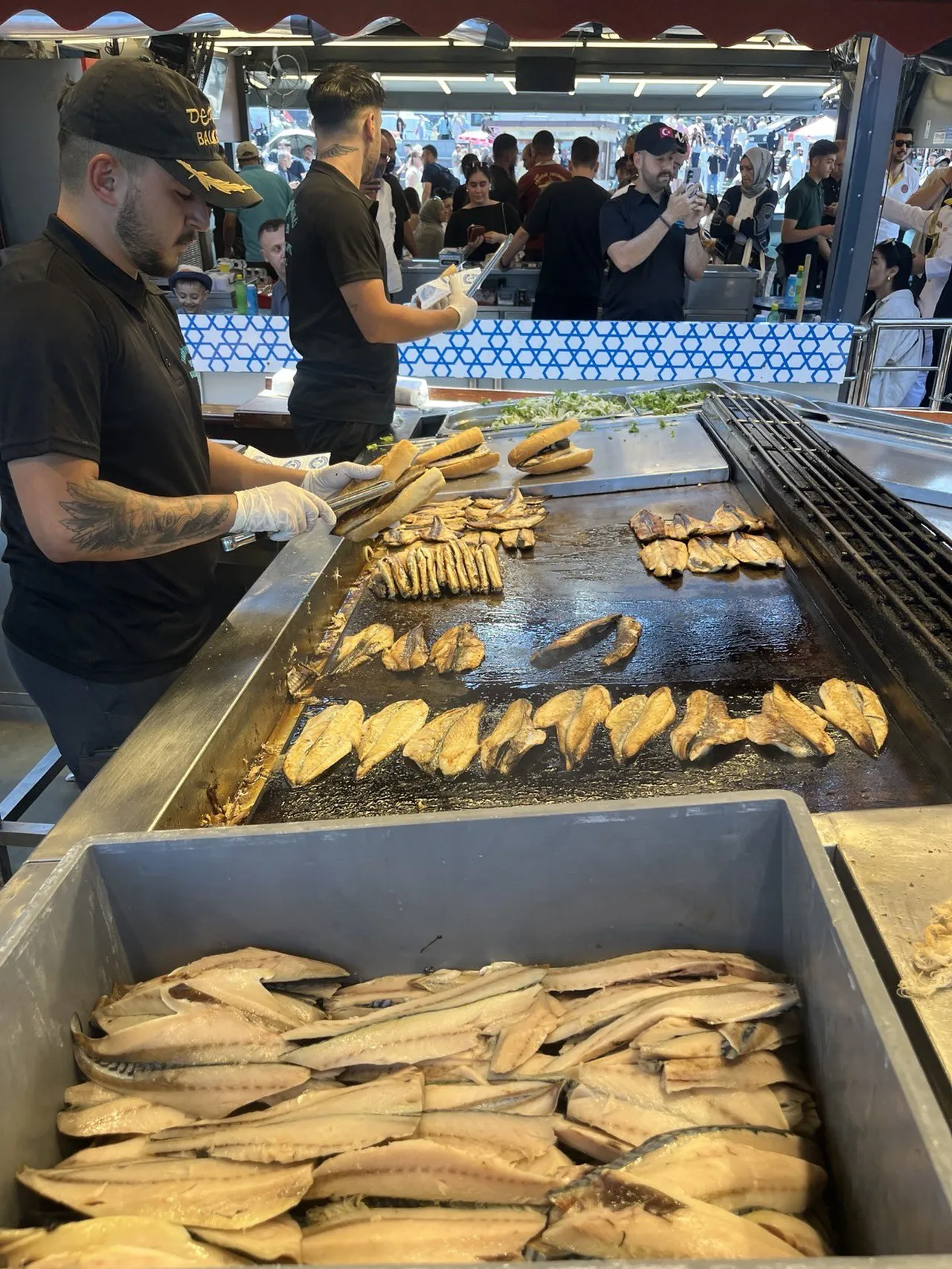 Kalabalık bir balık ekmek tezgahında ızgarada pişen balıklar ve sandviç hazırlayan çalışanlar.