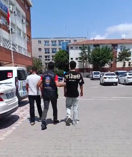 Polis eşliğinde yürüyen iki kişi ve emniyet binası çevresindeki araçlar.