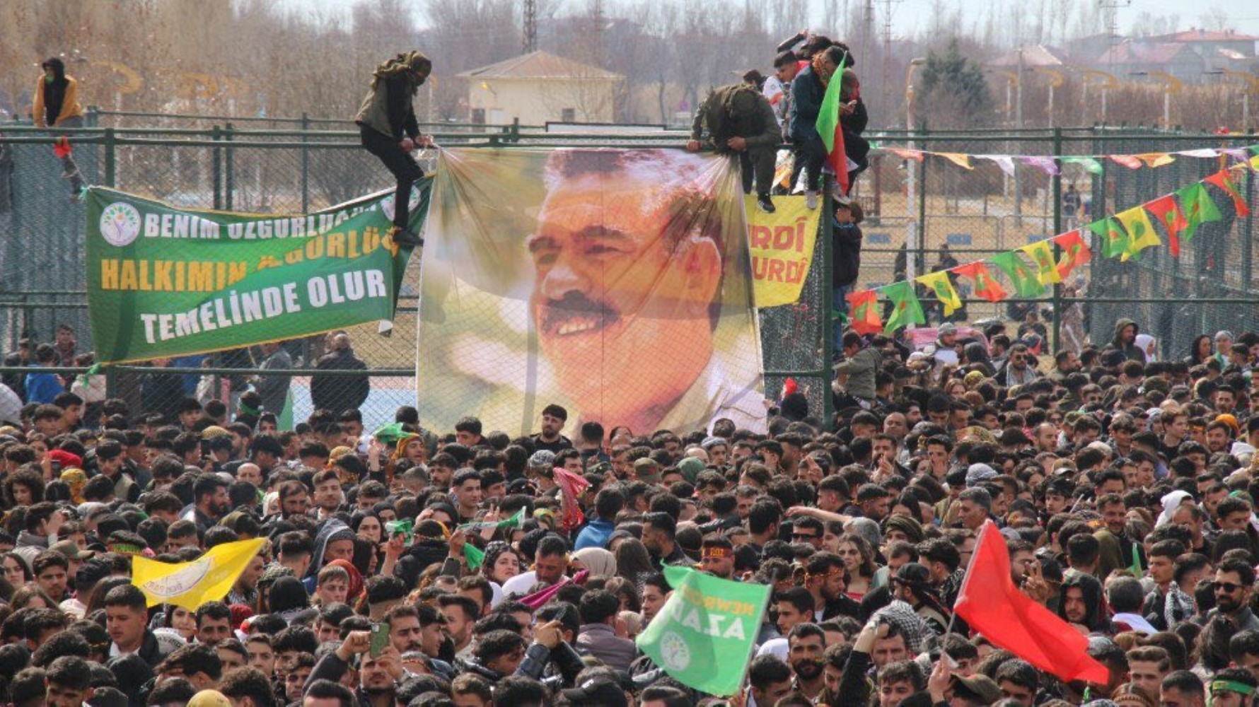 Kalabalık bir alanda büyük bir posterin asıldığı ve çok sayıda kişinin toplandığı etkinlik görülüyor.