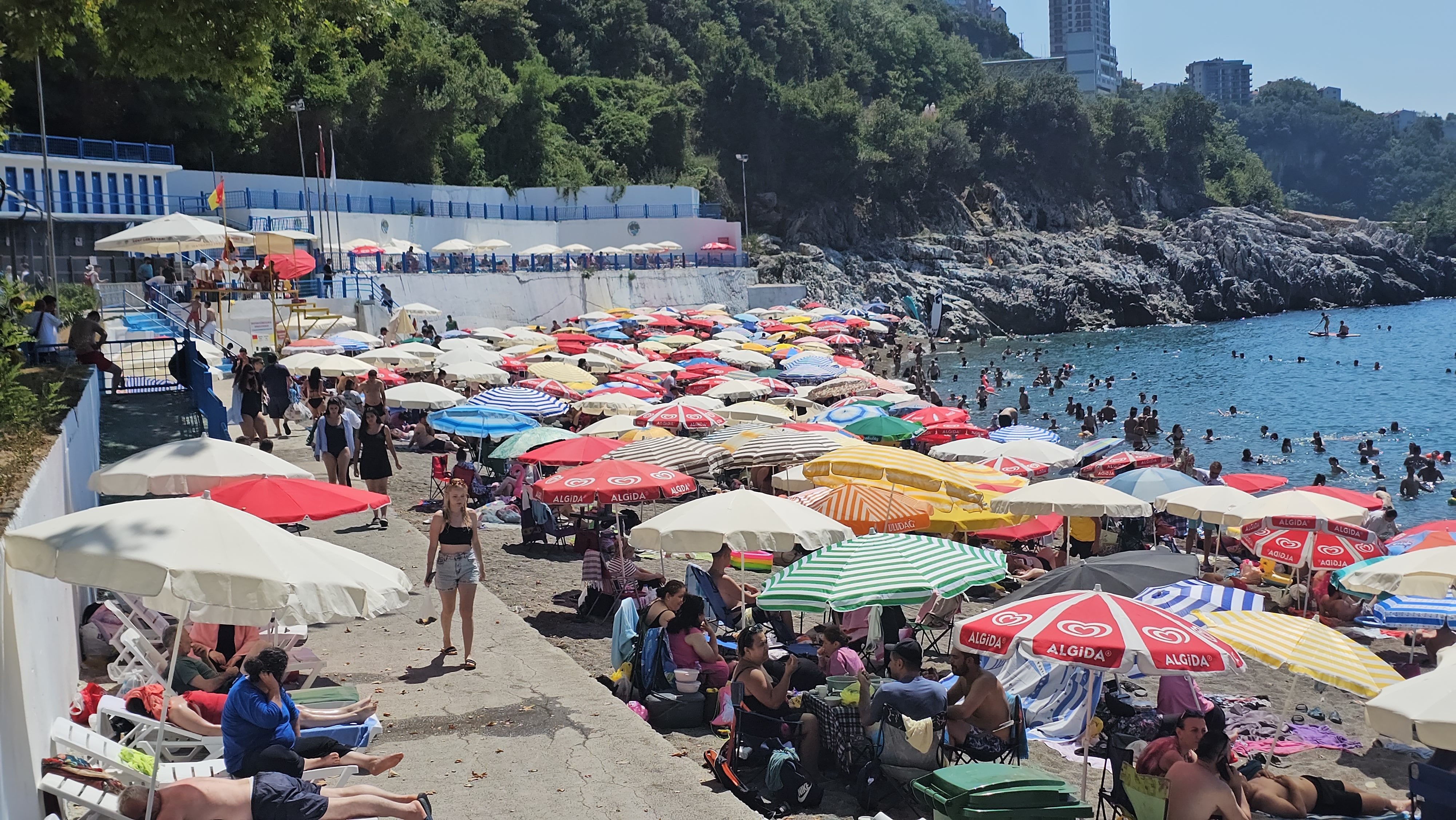 Kapuz Plajı’nda şemsiyelerle dolu kumsal, yürüyen tatilciler ve denize giren kalabalık.