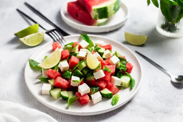 Beyaz masa örtüsü üzerinde karpuz, beyaz peynir, salatalık ve limon dilimleri içeren beyaz tabak.