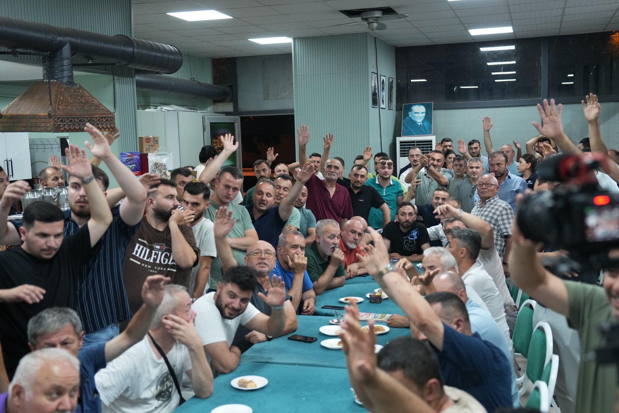 Kalabalık bir toplantı salonunda ellerini kaldıran katılımcıların görüldüğü fotoğraf.