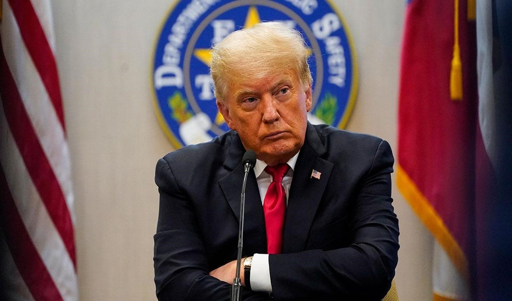 Donald Trump, toplantı salonunda mikrofon önünde otururken görülüyor.