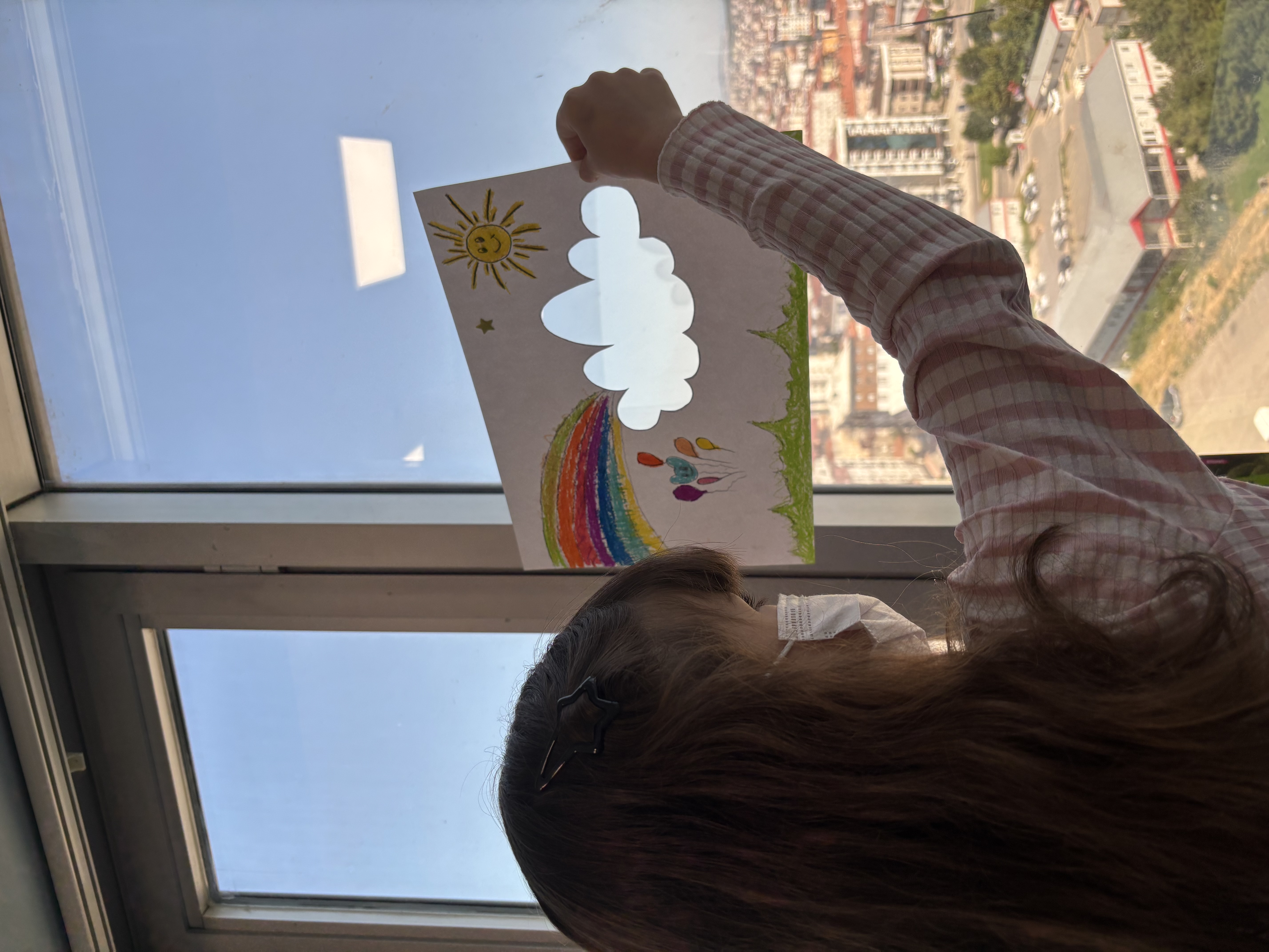Pencere önünde elindeki renkli çizimi tutan bir çocuk.