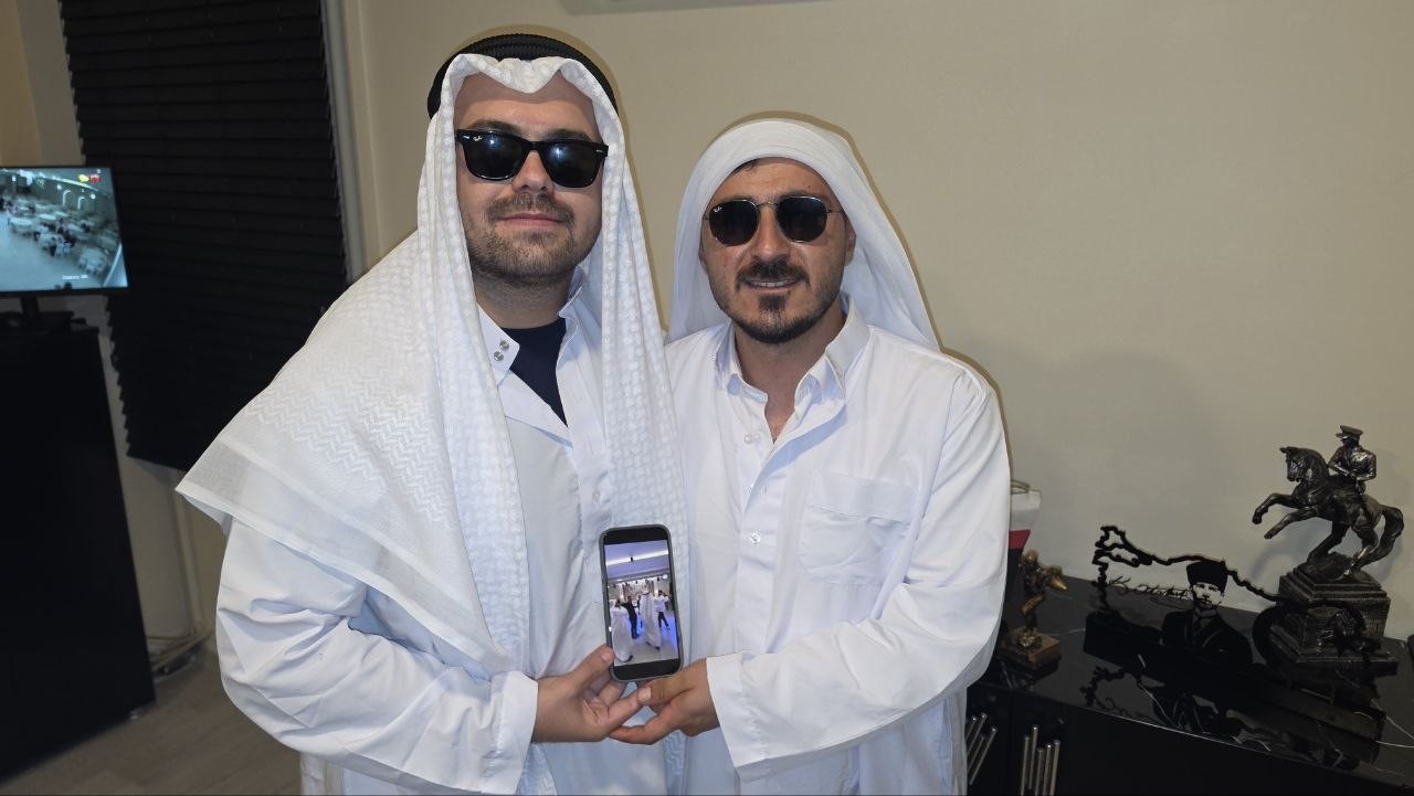 Arap kıyafetleri ve güneş gözlükleri takan iki kişi, bir telefon ekranını kameraya göstererek poz veriyor.