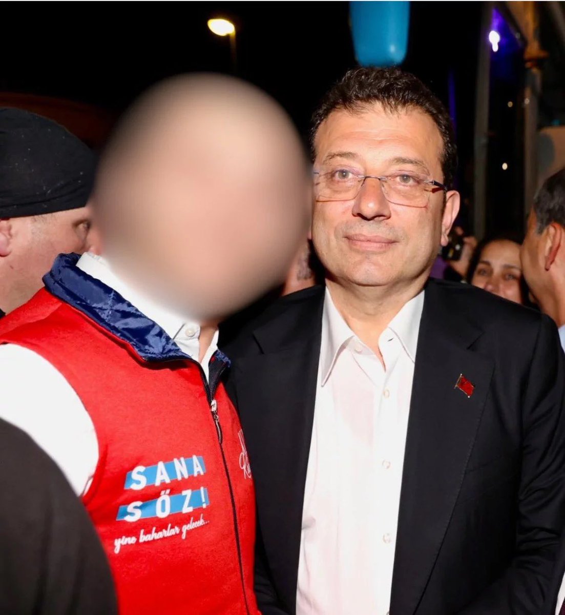 Gece çekilmiş, kalabalık içinde iki kişinin görüldüğü ve bir yüzün bulanıklaştırıldığı fotoğraf.