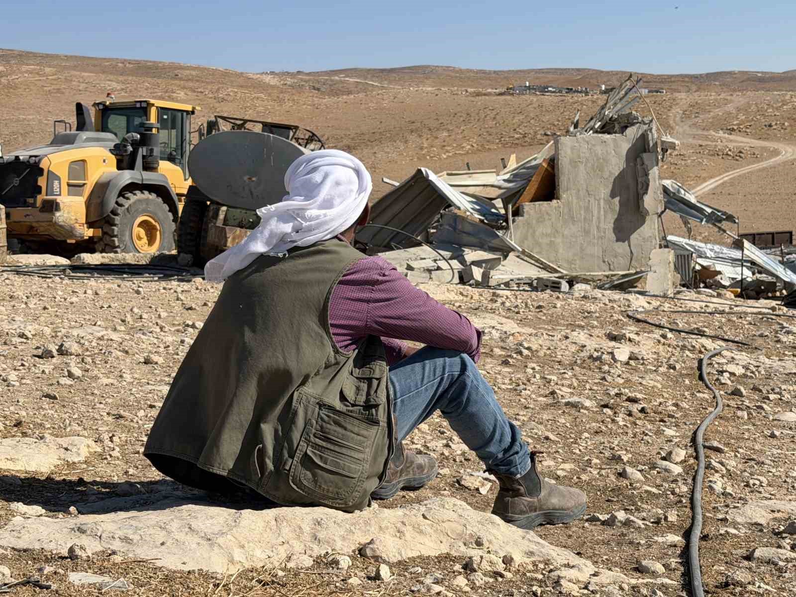 Bir kişinin taşlık arazide oturarak yıkılmış yapıya ve iş makinesine baktığı görüntü.