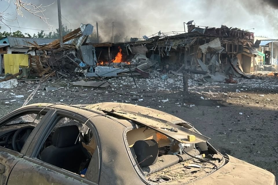 Hasar gören bir araç, yıkılmış yapılar ve arka planda devam eden yangın görülüyor.