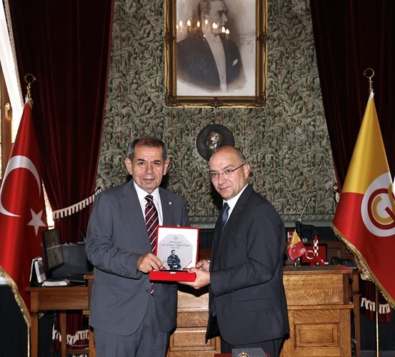 Galatasaray Lisesi’nde düzenlenen törende iki kişi anı plaketiyle poz veriyor; arka planda Türk bayrağı ve Galatasaray bayrağı bulunuyor.