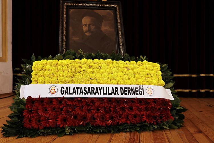 Tevfik Fikret portresi önünde Galatasaraylılar Derneği yazılı kırmızı ve sarı çiçeklerden oluşan çelenk.