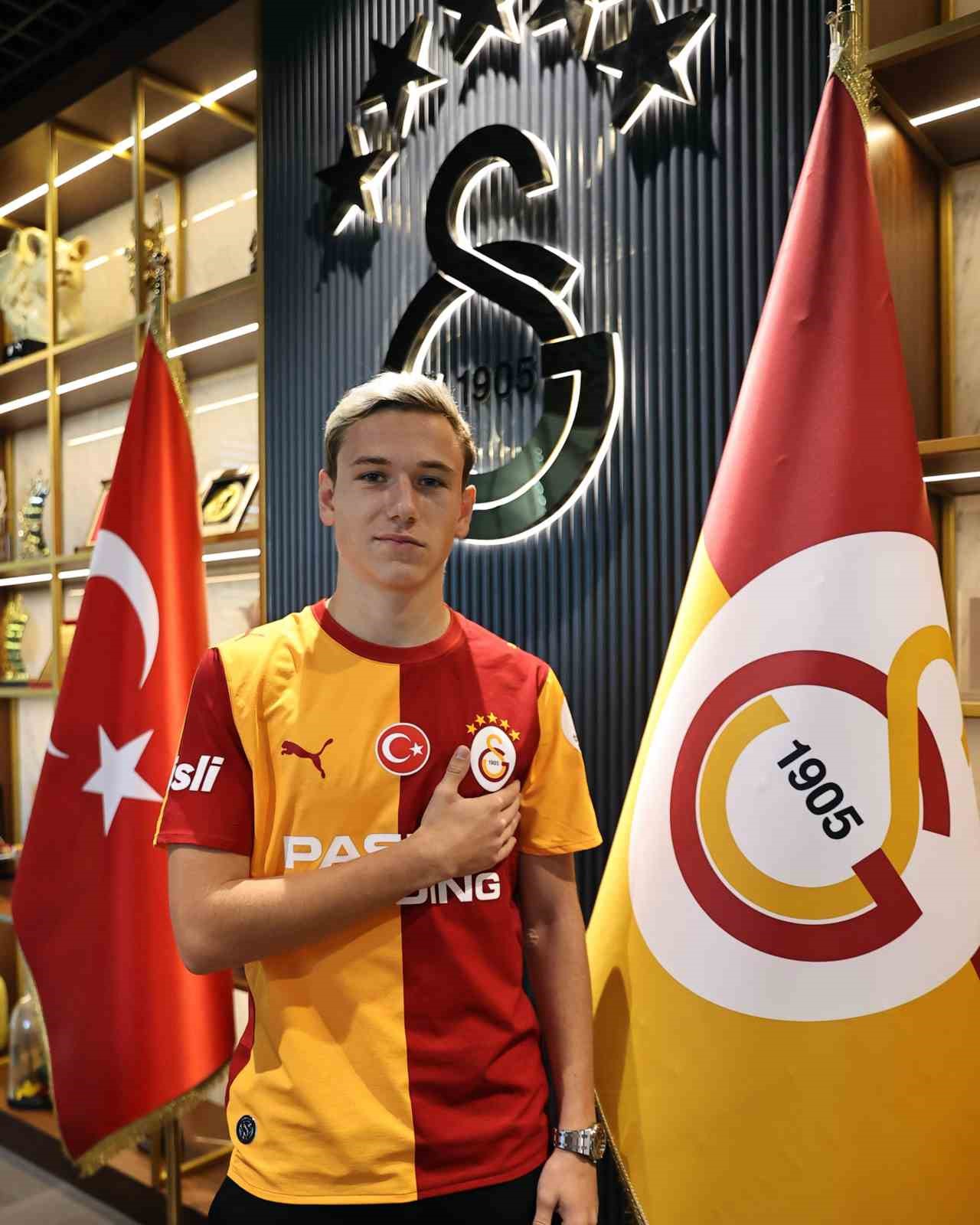 Aleksey Batrakov, Galatasaray bayrakları ve kulüp logosu önünde forma ile poz veriyor.