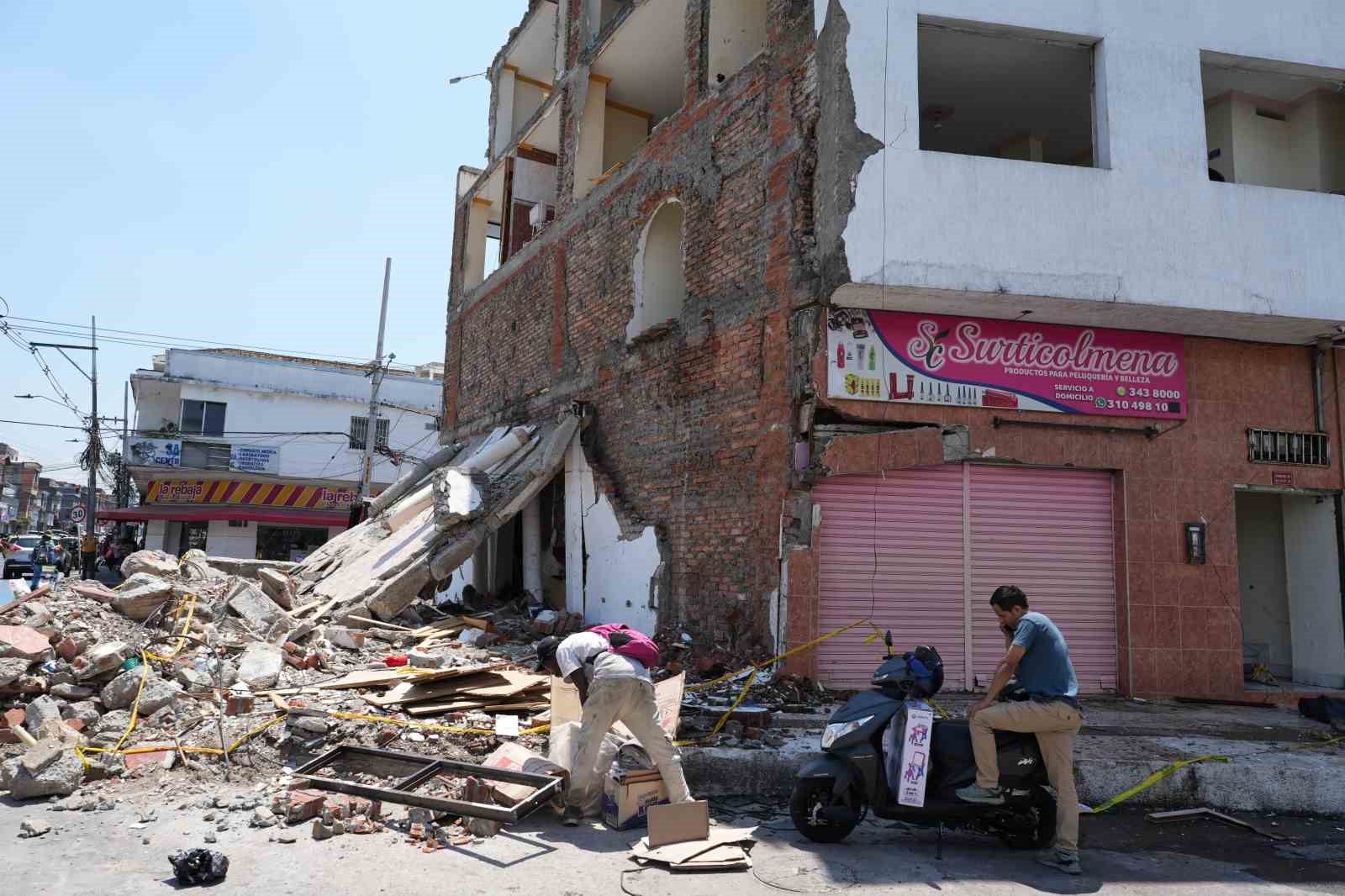 Bir sokakta deprem nedeniyle kısmen yıkılmış bina, molozlar ve çevrede bulunan kişiler.