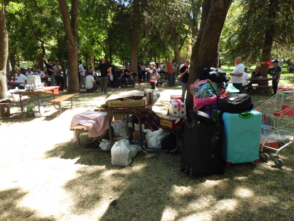 Kurtuluş Parkı’nda masaların yanında valizler, çantalar ve çeşitli ihtiyaç malzemeleri.