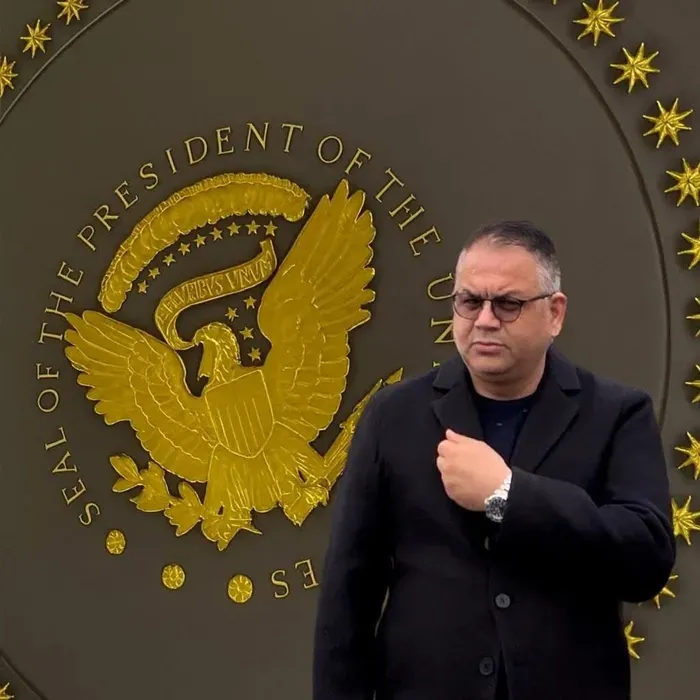 Bir erkeğin, üzerinde ABD Başkanlığı mührünü andıran büyük bir amblemin bulunduğu duvar önünde durduğu görsel.