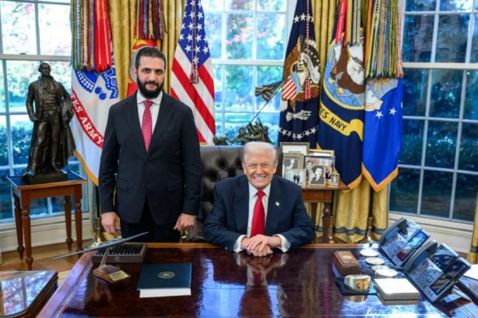 ABD bayraklarının bulunduğu resmi bir odada Donald Trump ve yanında ayakta duran bir kişi görülüyor.