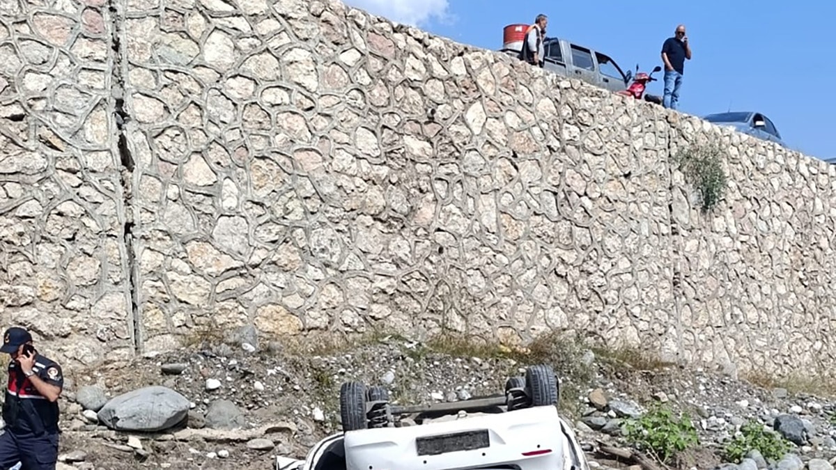 Dereye uçan otomobilin bulunduğu alanda taş duvar, yol kenarı ve inceleme yapan görevliler.