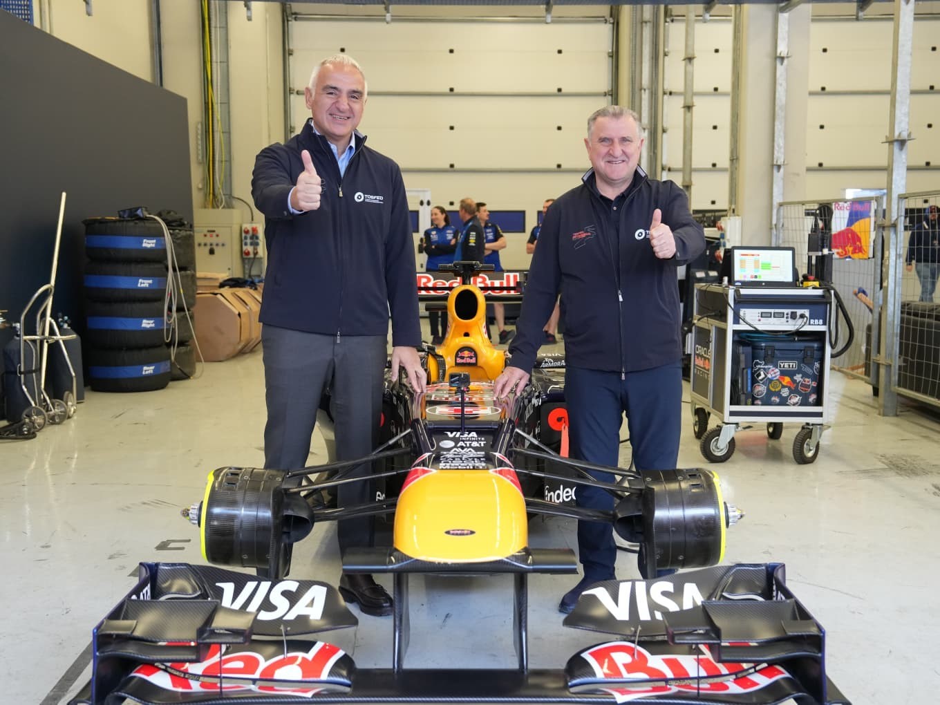 Garaj alanında Formula 1 aracının yanında poz veren iki yetkili.