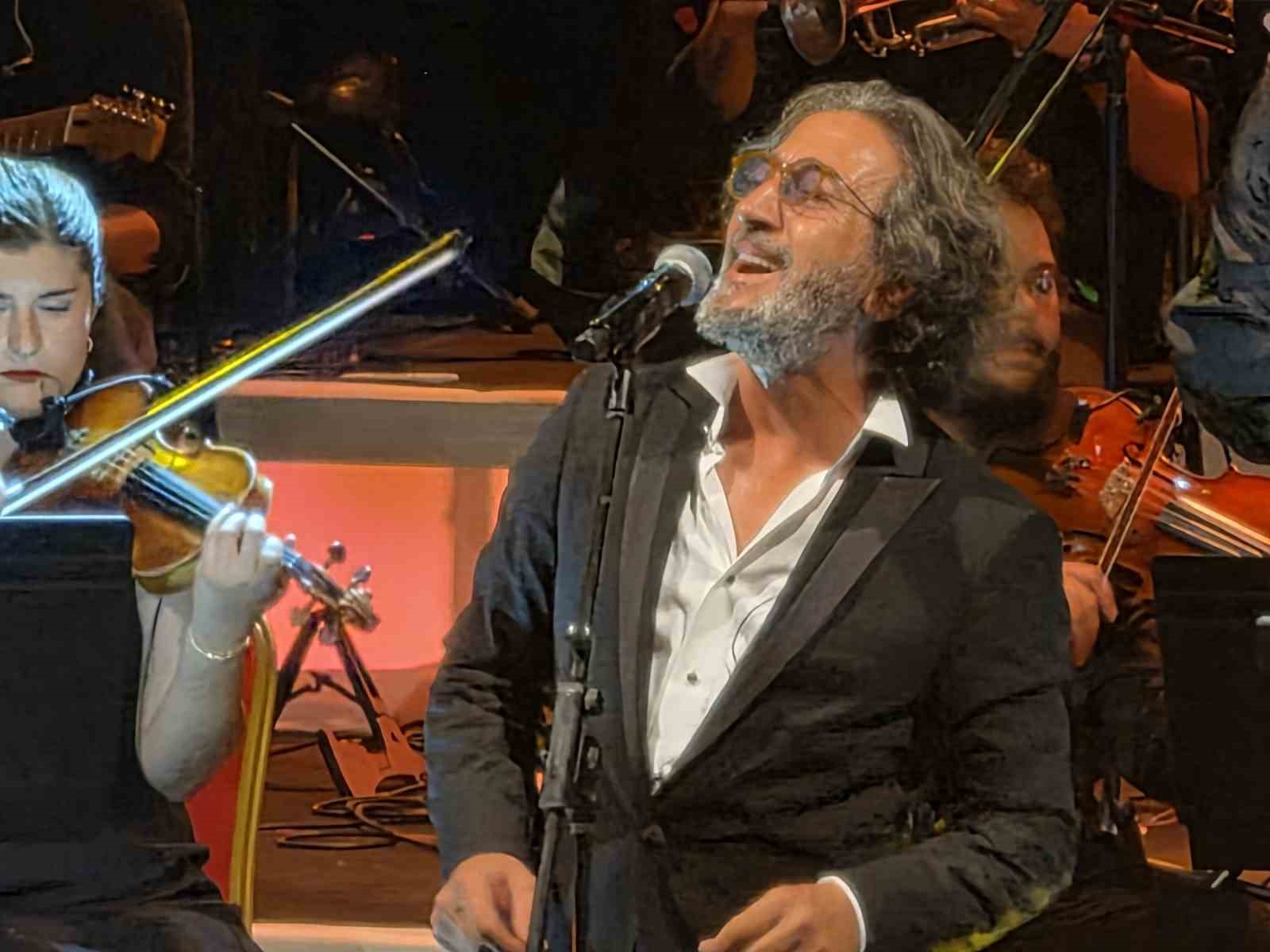 Fettah Can, orkestra eşliğinde sahnede şarkı söylerken yan profilden görülüyor.