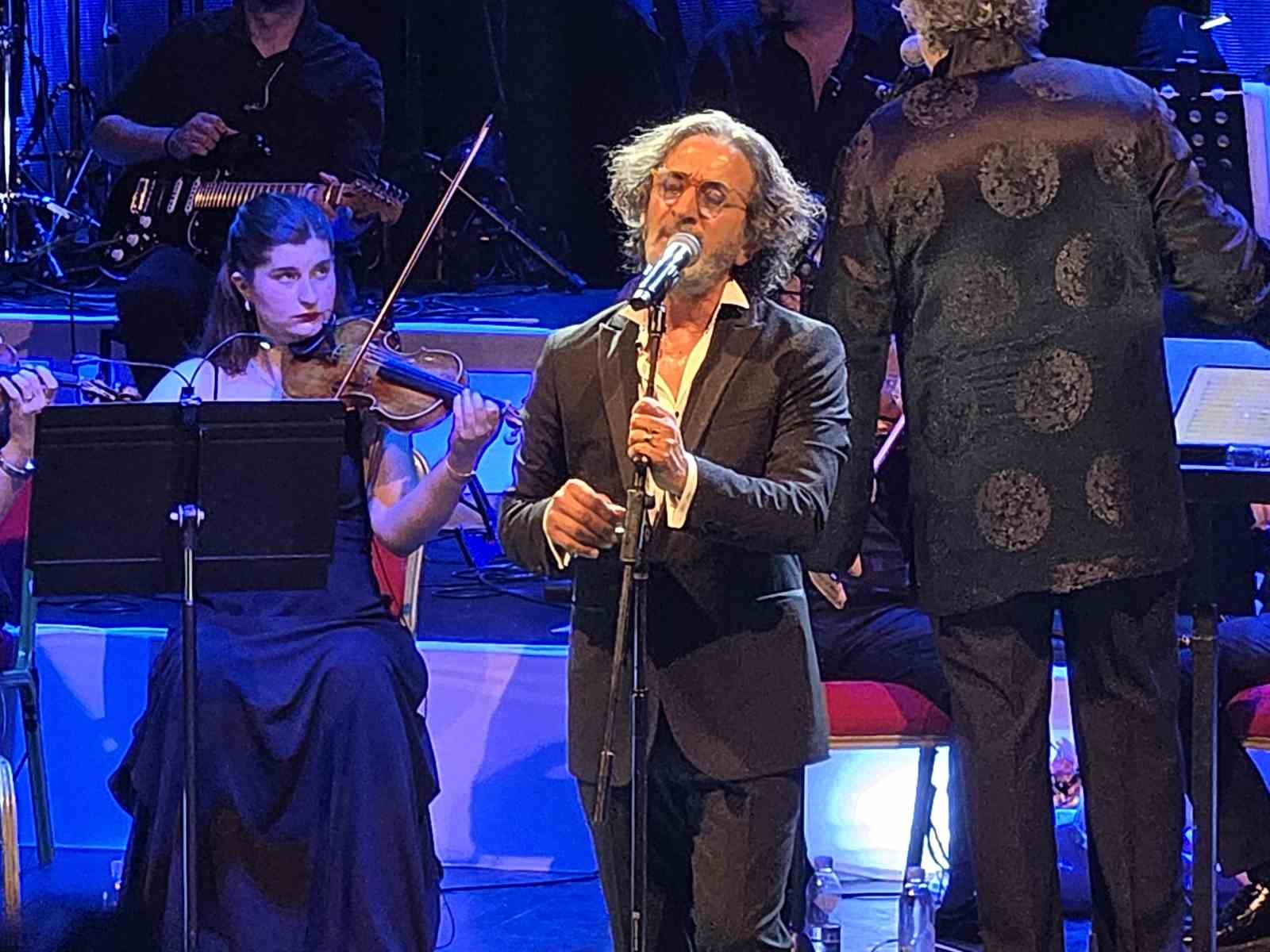 Fettah Can, mavi sahne ışıkları altında orkestra eşliğinde performans sergiliyor.