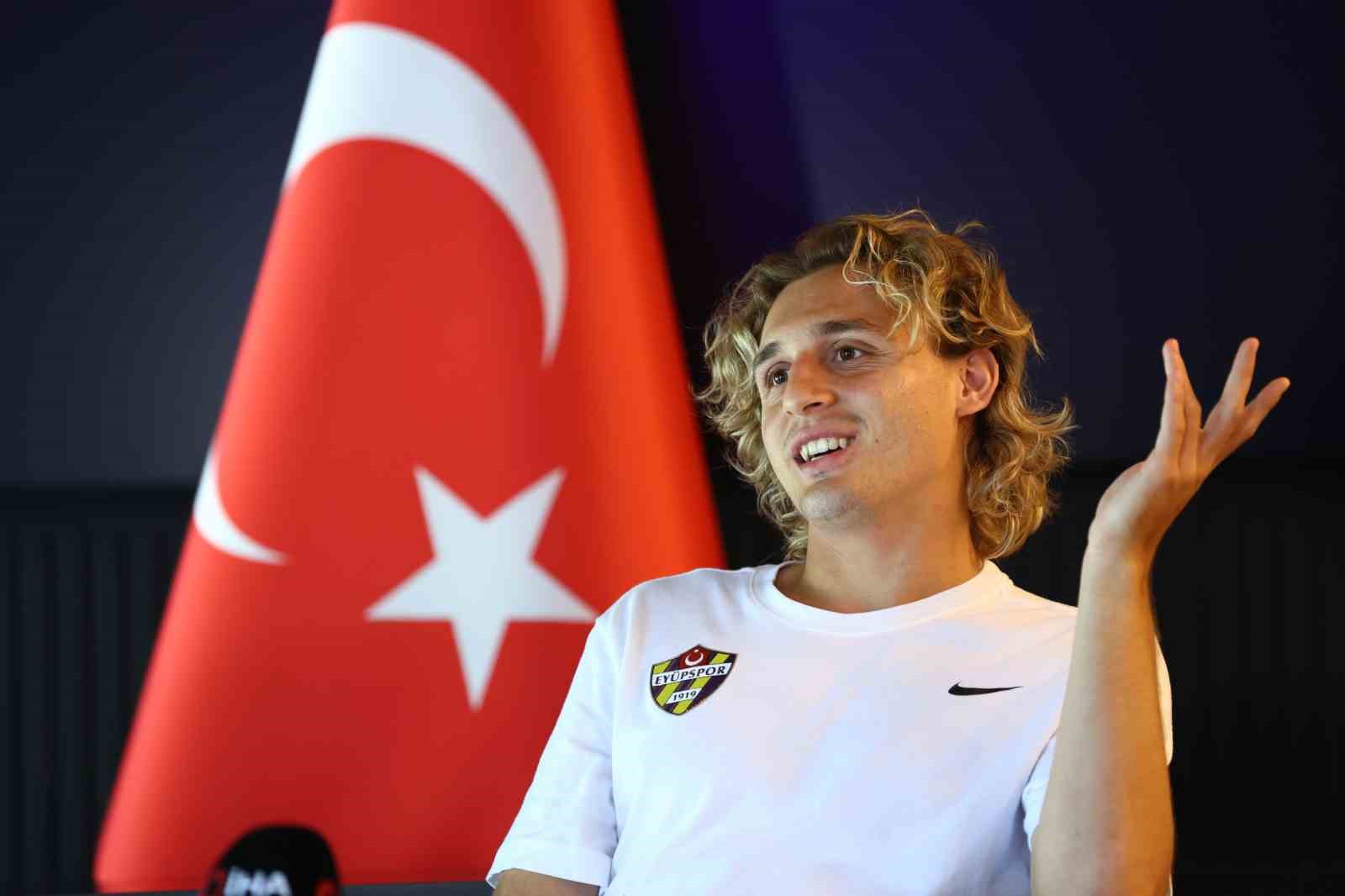 Hamza Akman, Türk bayrağı önünde röportaj sırasında jest yapıyor.